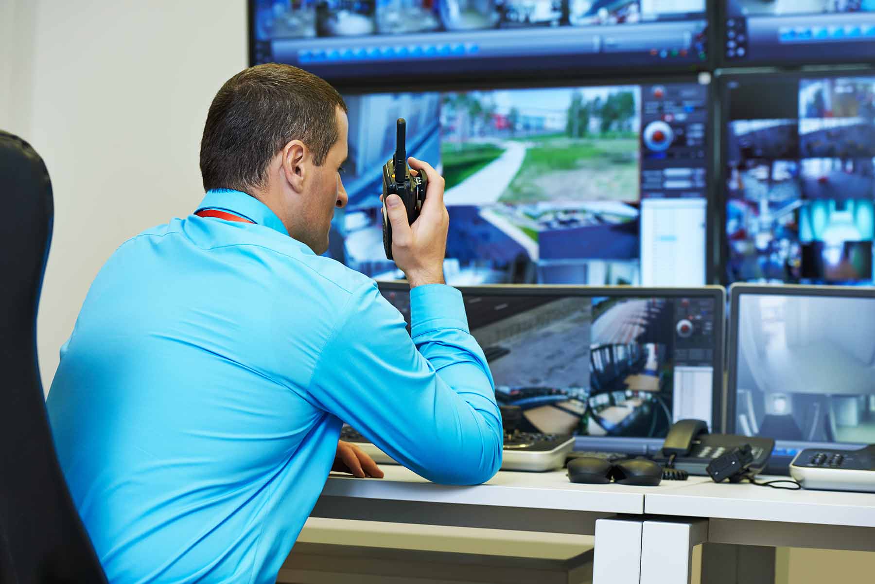 Security control room with a person monitoring multiple surveillance feeds while holding a walkie‑talkie.