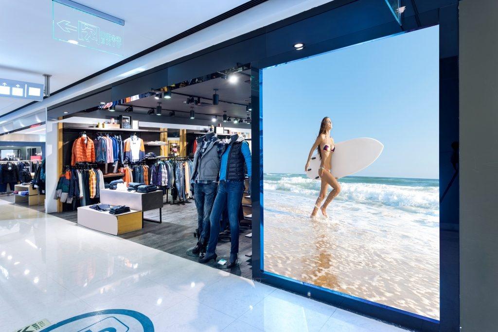 Clothing store entrance featuring mannequins in casual wear and a large digital display of a woman with a surfboard on the beach.