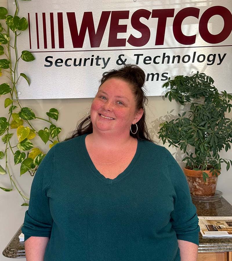 A smiling woman wearing a green V-neck sweater stands in front of a “WESTCO Security & Technology Systems” sign, with leafy potted plants in the background.