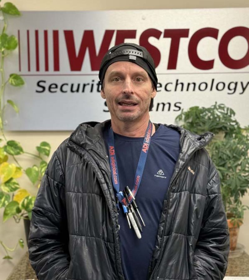 A man wearing a black puffer jacket, backward black cap, and a lanyard with keys stands in front of a “WESTCO Security & Technology Systems” sign, with green potted plants in the background.
