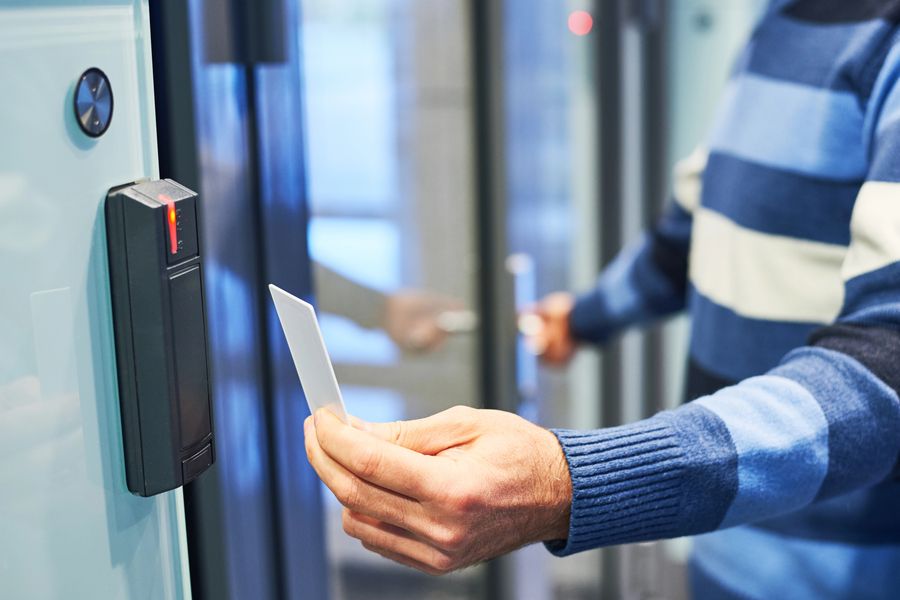 A person in a blue striped sweater holds a white keycard up to a black electronic keyless reader by a modern glass door.