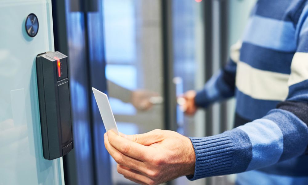 A person in a blue striped sweater holds a white keycard up to a black electronic keyless reader by a modern glass door.