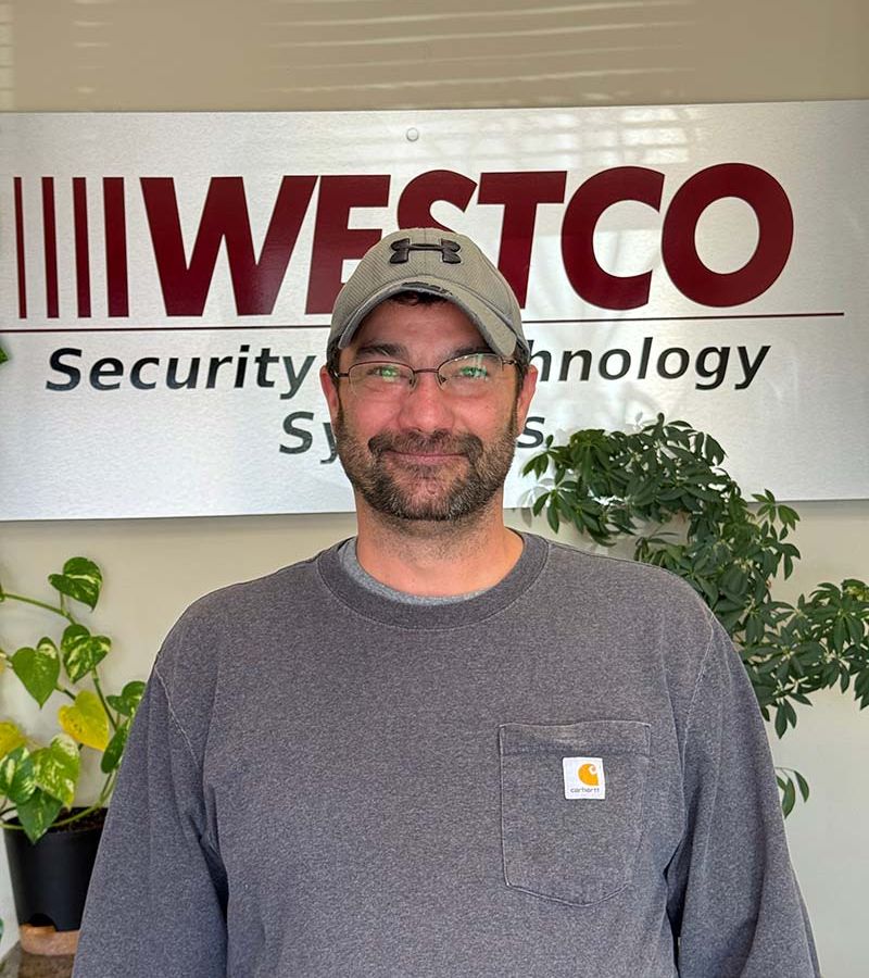 A smiling man wearing a gray Carhartt sweatshirt and a gray Under Armour baseball cap stands in front of a “WESTCO Security & Technology Systems” sign, with green potted plants in the background.