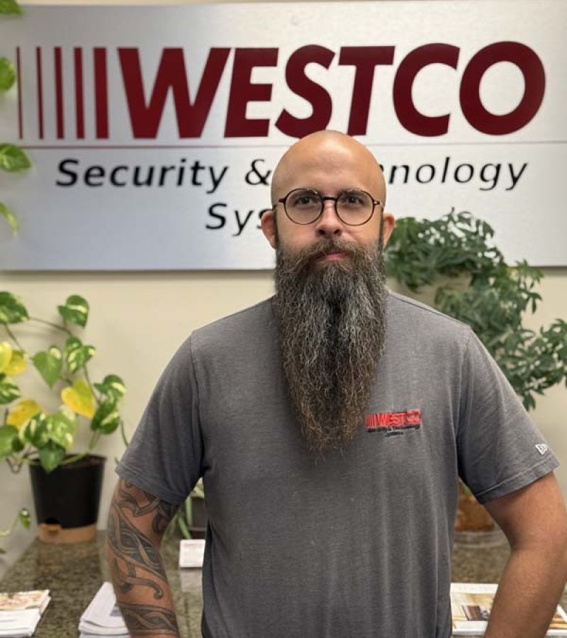 A bearded man with round glasses and tattooed arms stands in front of a “WESTCO Security & Technology Systems” sign, wearing a gray T-shirt with the Westco logo.