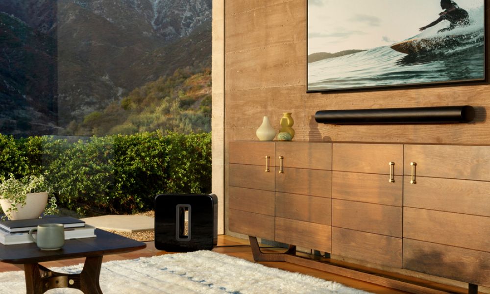 Black Sonos Arc soundbar mounted below a wall-mounted TV, with a Sonos Sub subwoofer on the floor nearby.