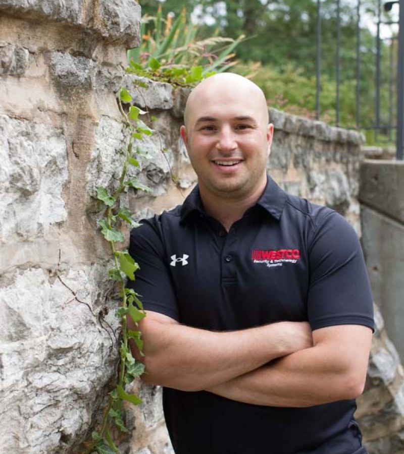A smiling man in a black “WESTCO” polo shirt stands with his arms crossed against a stone wall overgrown with ivy.