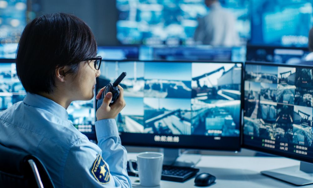 Security operator with a radio reviews camera feeds of an industrial yard on multiple monitors in a monitoring center.
