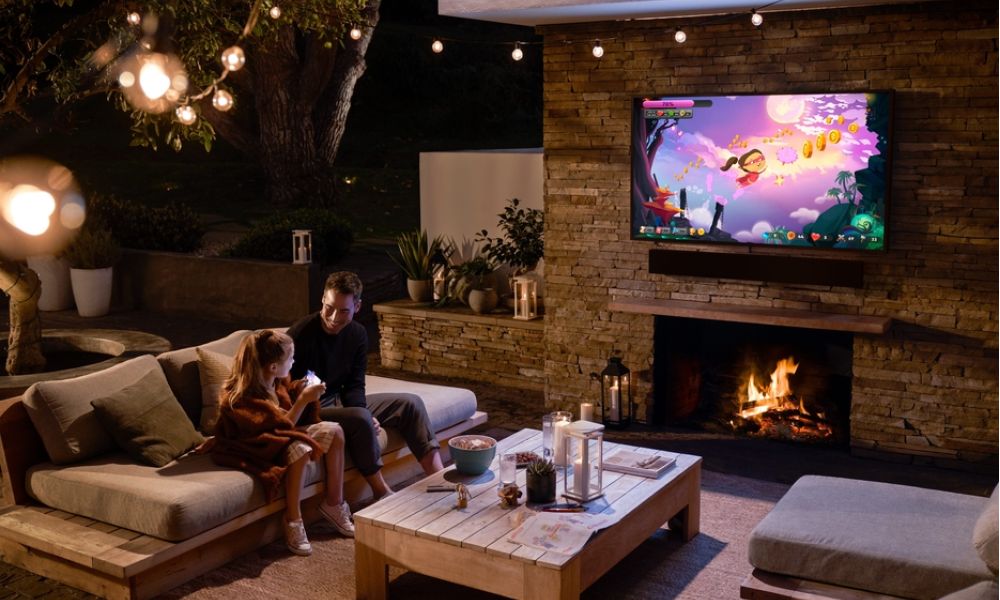 A man and young girl sit on an outdoor patio watching a video game on a wall-mounted TV above a fireplace.