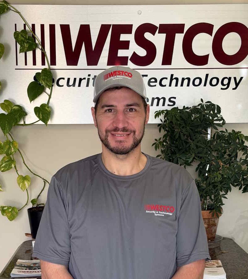 A man wearing a black puffer jacket, backward black cap, and a lanyard with keys stands in front of a “WESTCO Security & Technology Systems” sign, with green potted plants in the background.