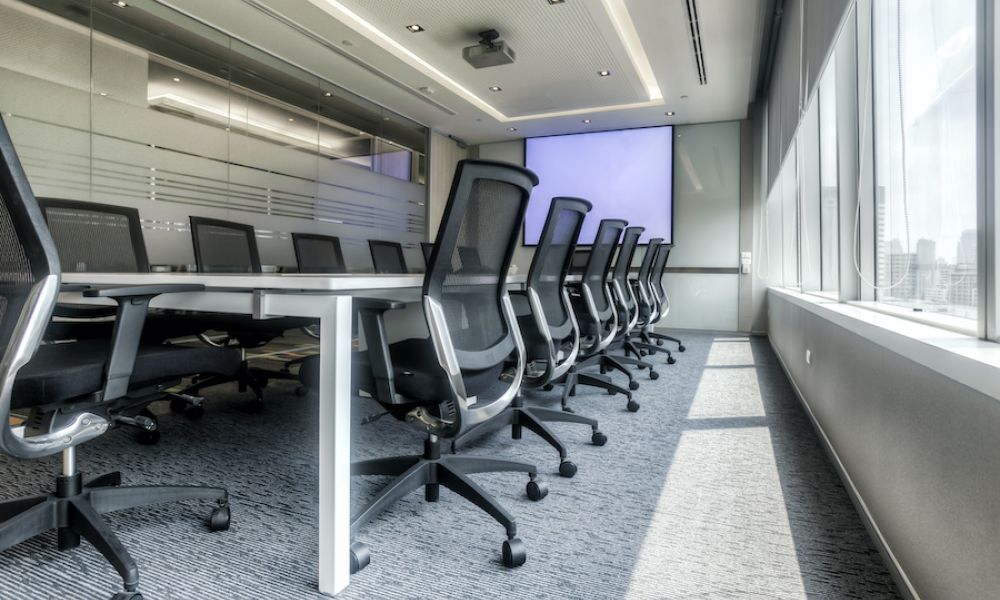 Bright, modern conference room with a long table lined by black mesh office chairs, large windows along one side, and a blank projection screen at the front.