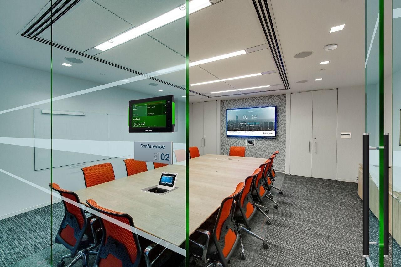 Modern conference room with a long table, orange chairs, wall-mounted displays, and a tablet integrated into the tabletop for meeting controls.