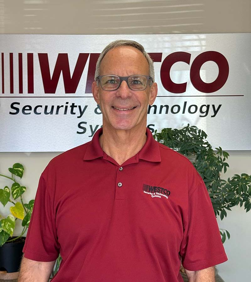 A smiling man in glasses wearing a red “WESTCO” polo shirt stands in front of a “WESTCO Security and Technology Systems” sign, with green potted plants behind him.