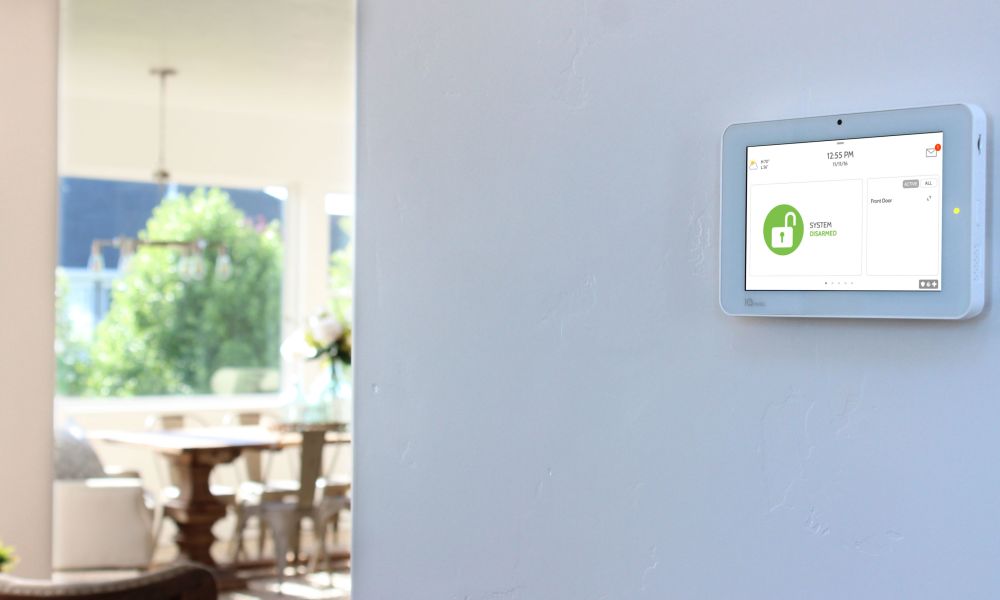 A home security touchscreen panel mounted on a beige wall near the front door, displaying a user interface.